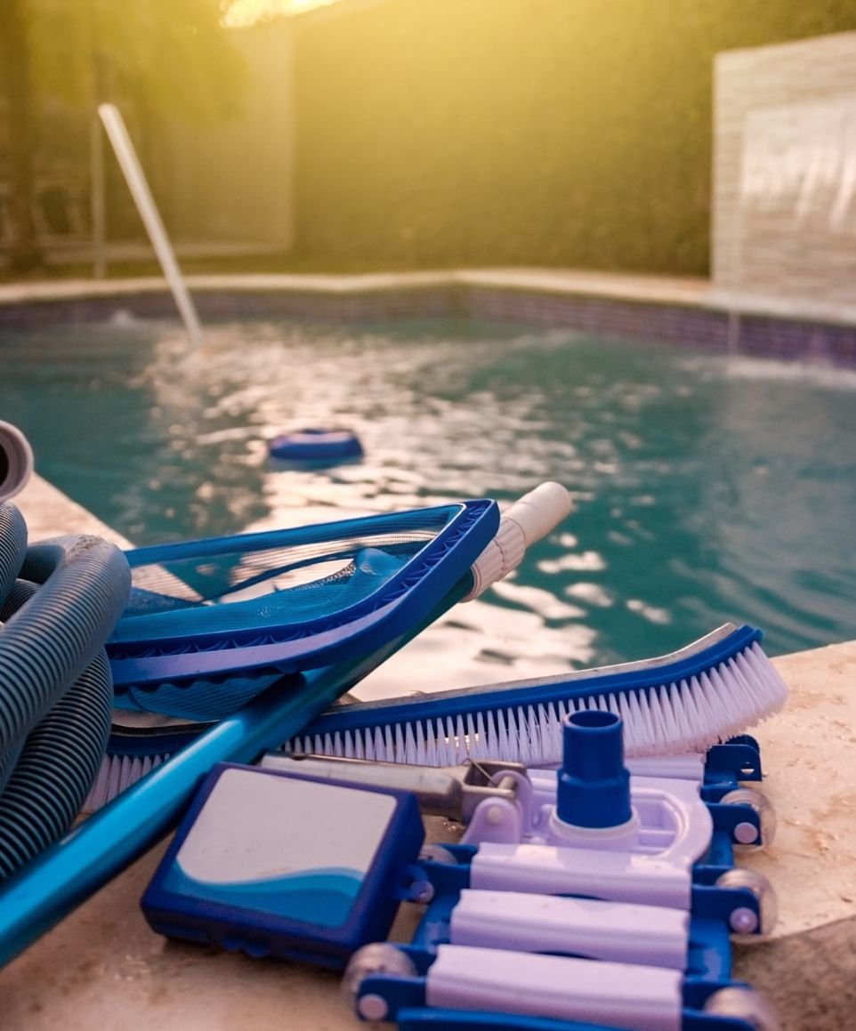 Pool cleaning equipment including brush, vacuum head, skimmer net, and hose placed beside a backyard swimming pool at sunset.